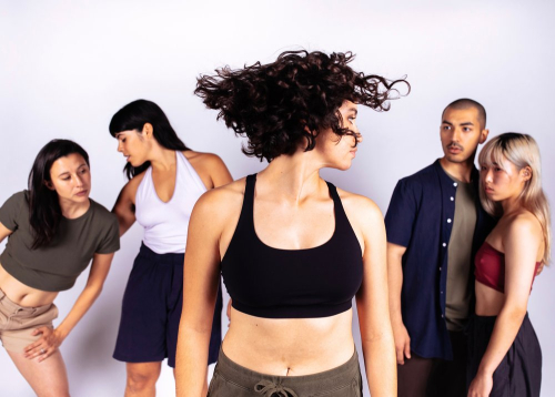 Five dancers pose. The dancer in the foreground turns her head to the left, hair flying in the air. Two sets of two dancers pose in the background.