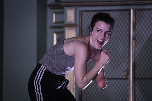 A dancer smiles to their right with open mouth. She wears a sparkly grey camisole top and black and white track pants. The photo captures here upper body and upper thighs.