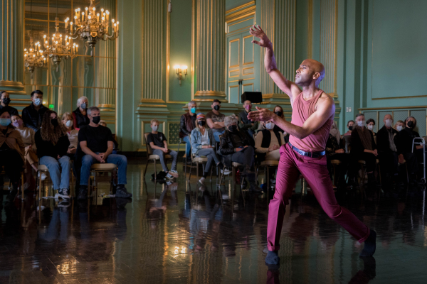 A solo dance poses with bent arms in the air, looking to his right. Audience members are seated behind him in the Green Room at the War Memorial building. Sparkly chandeliers and mint green walls.