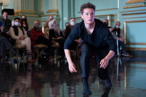 A solo dancer in a long sleeve black shirt and black pants and socks is captured in a photo lunging forward. Her focus and eyes are strongly forward. 