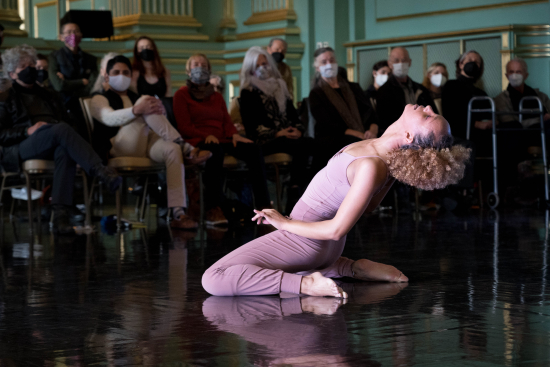 A dancer in dusty rose pink costume sits on her knees on the floor and leans back. You see the audience in front of her, watching with masks on.