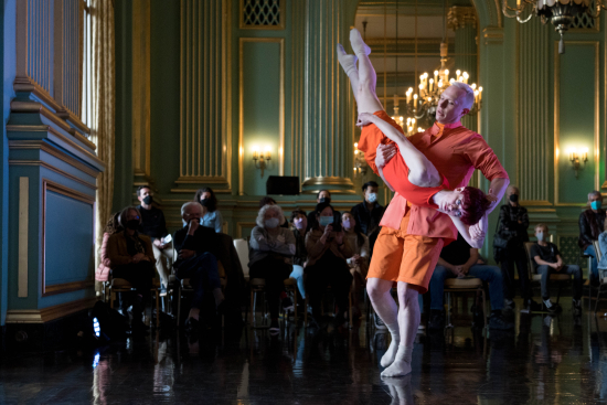 Two dancers are in orange attire. One dancer is lifted into the air, legs long and feet upward towards the ceiling.