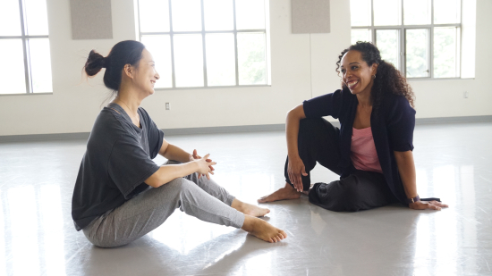 Two dancers sit and talk on the floor. The floor is a light grey marley, and the back wall is white with big windows.