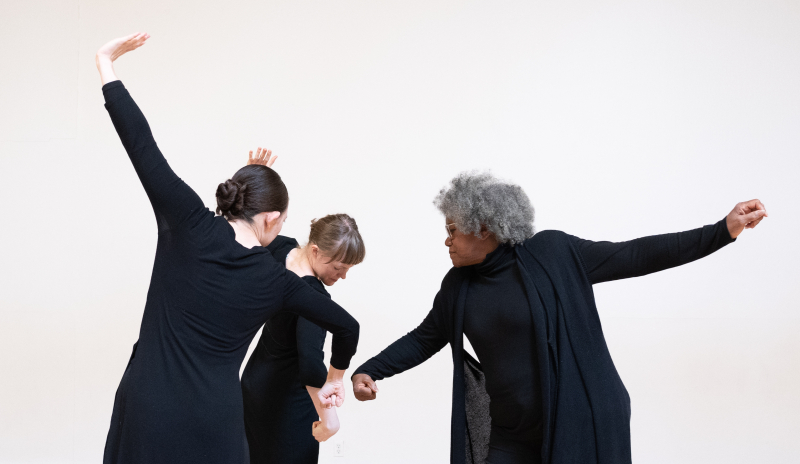 Three dancers wear all black in front of a white wall. They make a low fisted gesture towards the center of the circle.