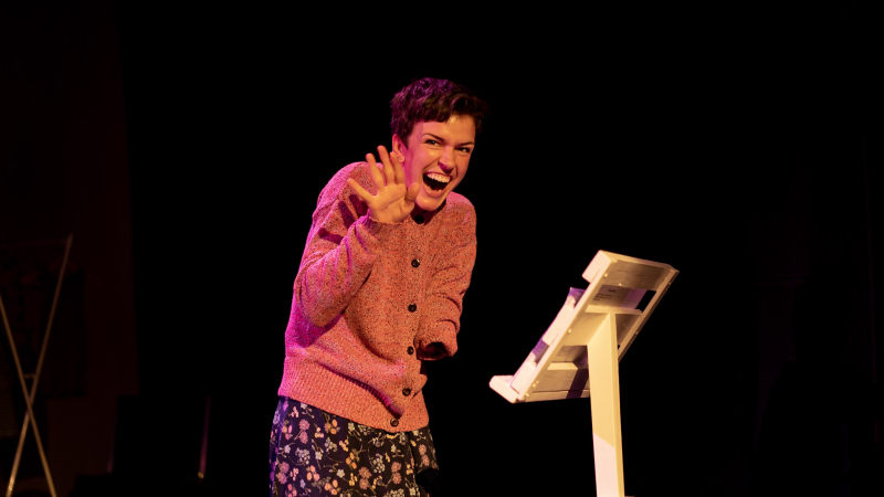 Julie makes a waving/hello gesture with her right hand and offers a large grin to the audience. She stands by a podium and wears a buttoned up orange/pink cardigan and long floral skirt. The background is dark.