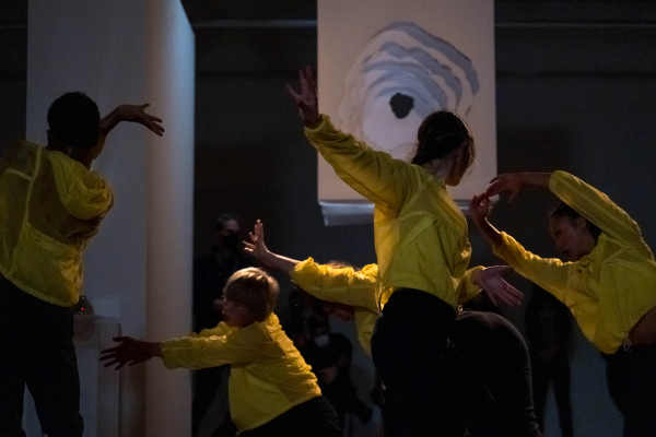 Five dancers wearing semi-translucent yellow jackets extend their upper limbs into various levels of space underneath a white hanging 3d paper sculpture with layers that create a deep tunnel. 