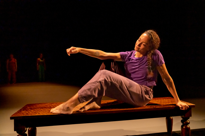  Shareen sits with two feet and one hand on a wooden bench, while her second hand gestures in front of her.