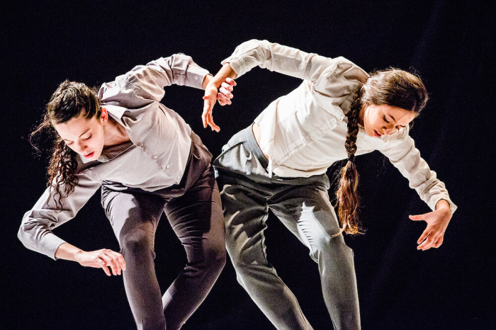 Two dancers from Vertigo Dance Company bending over and looking down in opposite directions.