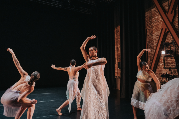 Five dancers in white flowy costumes dance downstage at ODC theater. They are captured posing in different shapes.