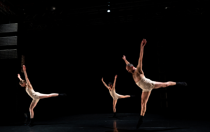 A trio of dancers in white costumes in front of a black, dark background. The dancers are caught in motion with arms up in a high V, their left leg back behind them in an arabesque.