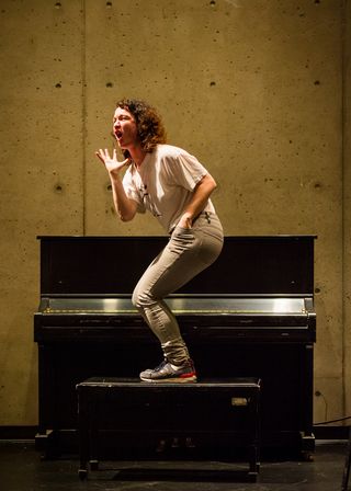 A dancer stands on a piano bench next to a black upright piano.