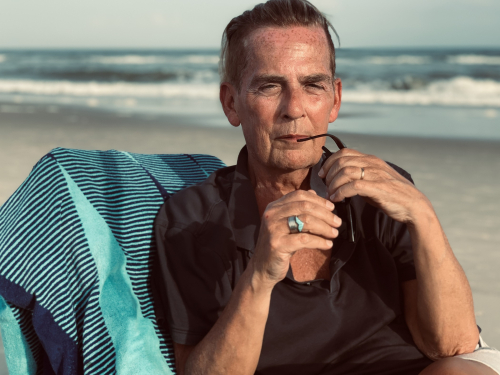 A photo of a man (upper body) sitting at the beach. He wears a brown short sleeved shirt and sits on a cair with a blue beach towel draped on it.