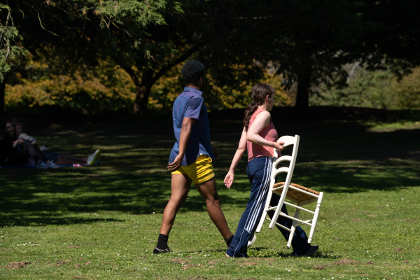 Two dancers walking, with one holding a chair.