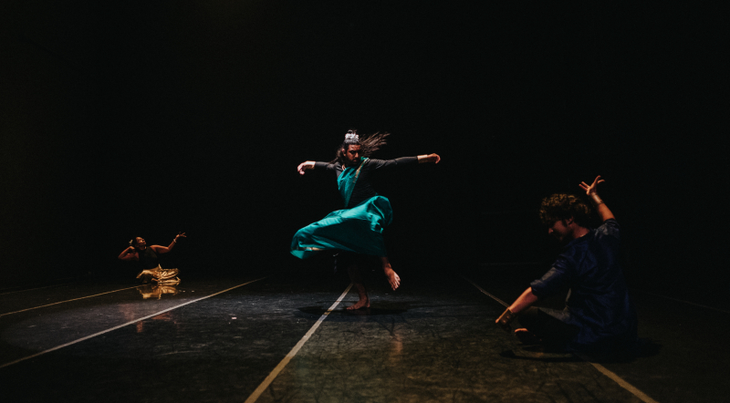 Trio of dancers performing in a dark space with black walls and black marley flooring. A dancer spins in the center with emerald green costume flowing. The other two dancers pose on the floor, one downstage right and one upstage left.
