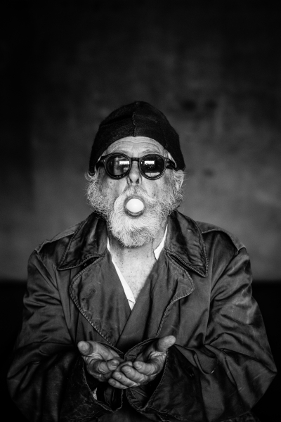 A man with fair skin, gray hair, and a beard is facing the camera in this black and white photo. He is wearing a trench coat, black knit cap, dark sunglasses and has an egg in his mouth. His arms are reaching out in front of him with his hands together as if at communion.