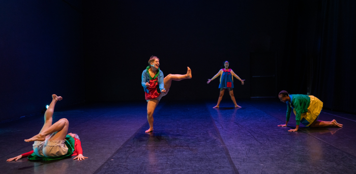 Four dancers pose in playful shapes on the floor and standing, wearing multicolor bright costumes.