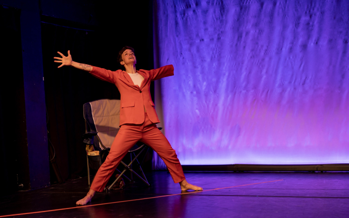 Julie makes a large X-shape pose. She wears a red blazer with matching red pants. The backdrop is purple and pink. Julie makes a large X-shape pose. She wears a red blazer with matching red pants. The backdrop is purple and pink.
