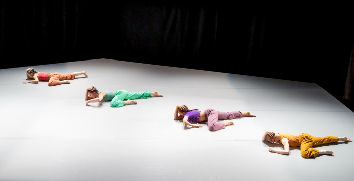 4 dancers, each wearing a different color, lay in nearly identical positions on a stark floor.