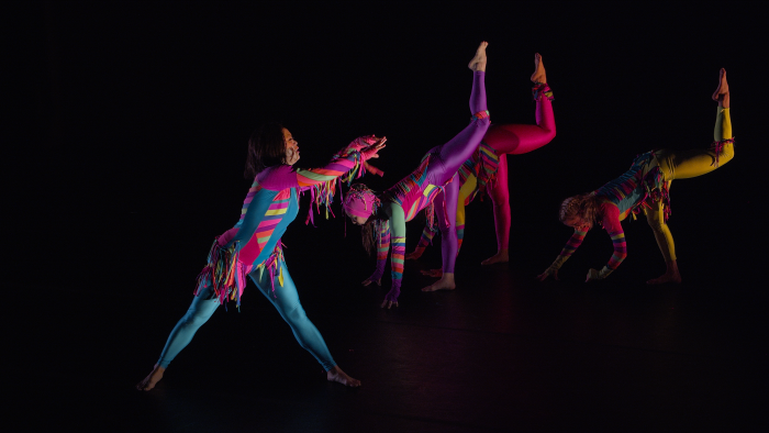 Four dancers in bright colors. One stands with feet wide apart in front of the group. The other three dancers are upstage, pitched upside down with hands on the ground and left leg in the air.
