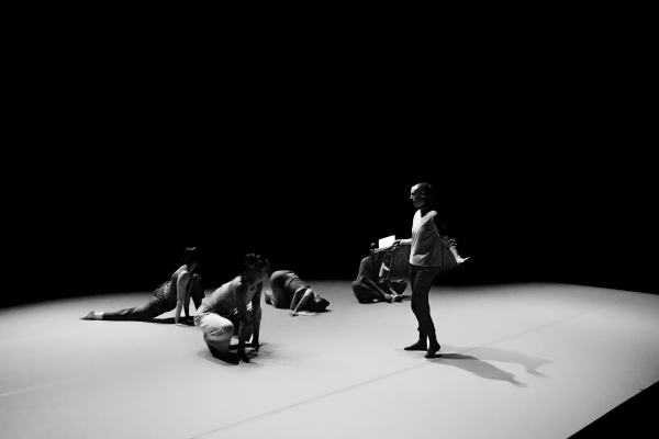 A black and white image of a person standing with a notebook surrounded by dancers in various stretches.