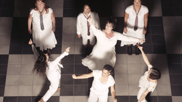 Three dancers standing and looking up at the camera; four dancers moving through the space dressed in white. 