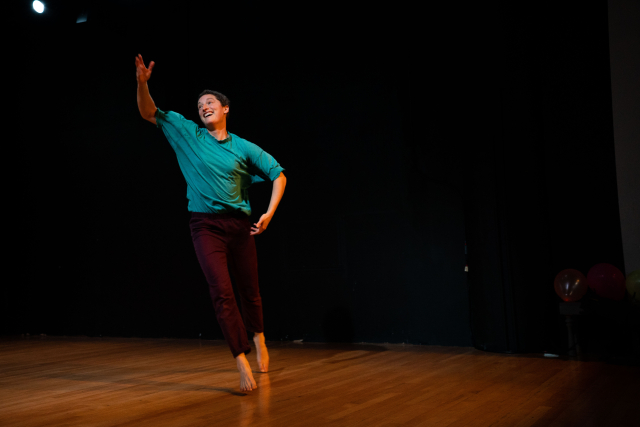 Dancer caught in motion, smiling at the audience. Her right arm is bent and her right hand is gesturing upwards towards the ceiling.