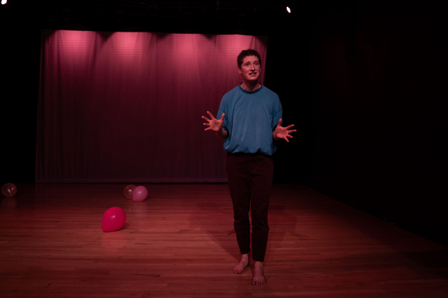 Dancer talks to audience and has palms by waist, facing audience. Balloons are strewn on the floor.