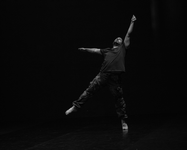 A black and white photo of Andrew Merrell. Background is dark/black. He poses with one arm above his head, one arm out to the side. One leg is reaching off of the ground.