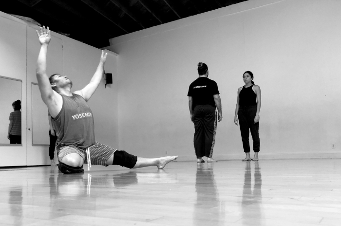 A black and white image of 3 dancers. The dancer in the foreground is on the floor, with his arms up in a V and is looking up.