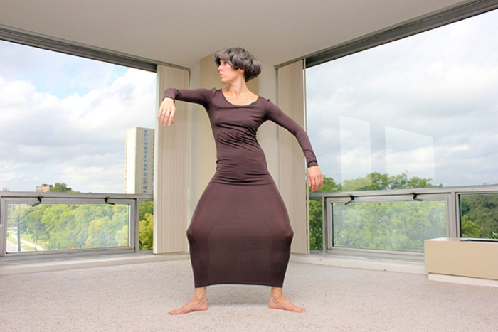 A dancer in a long, form-fitting brown dress poses and looks to their right.