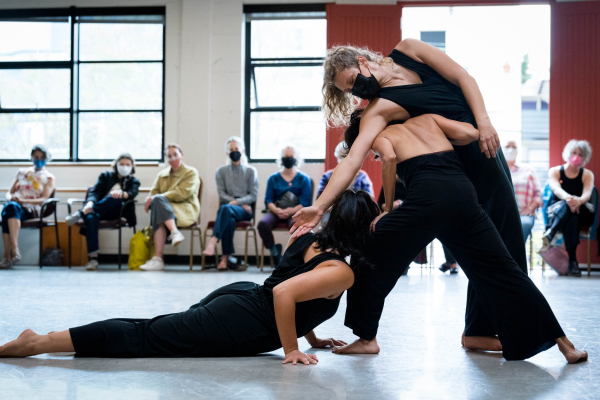 Three dancers in black costumes pose. One is on the floor. and one supports another dancer, leaning on her upper back.