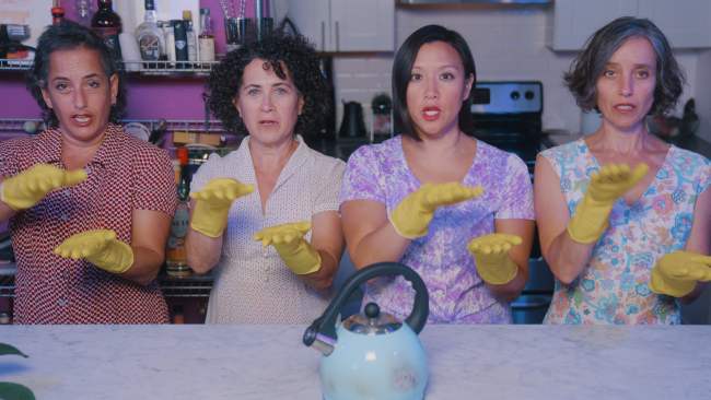 Four women wear dresses and yellow latex gloves. We see them from the waist up in front of a kitchen counter, with a blue tea kettle on it.