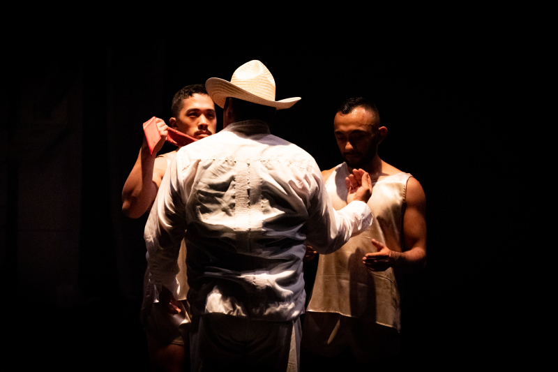 Two dancers help a third dancer get dressed in a Veracruz style folklorico costume with white shirt and pants and a hat.