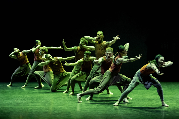 Ten dancers in a line pose with strained facial expressions, open mouths, and one arm reaching out.