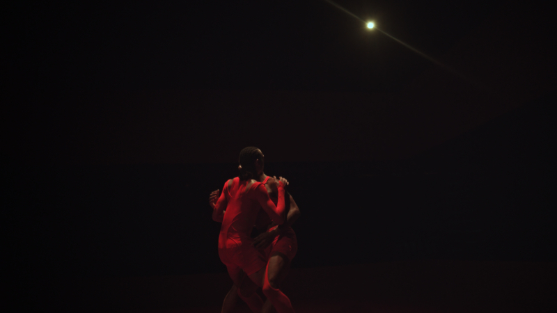 Two dancers in red costumes dance in front of a black background.