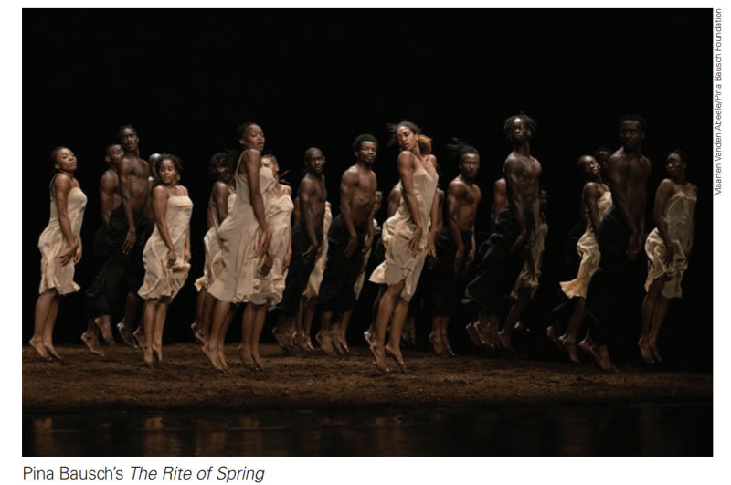 A stage full of dancers in dirt. The women wear cream silk long slips. The men wear black pants and no tops.
