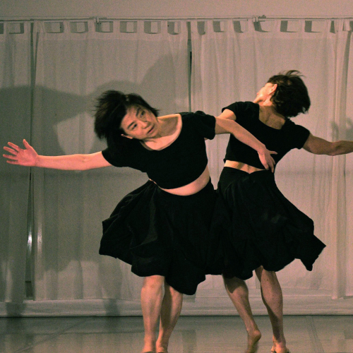 Two dancers spin together in black crop shirts and flowing knee length black skirts.