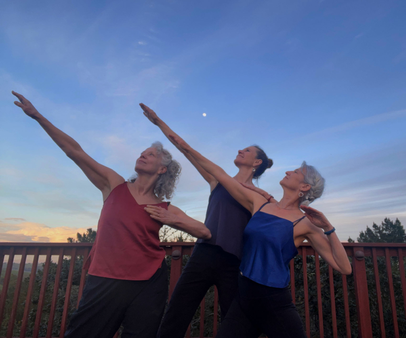 Three dancers on a deck pose with their right arms high at a angle.