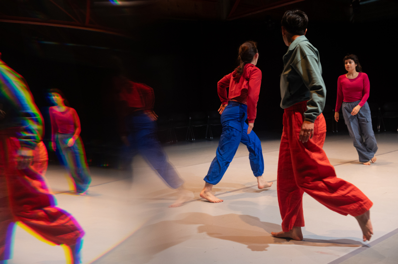 Three dancers, wearing blues, oranges, and pinks, are caught walking in different directions. A mirror effect on the image causes blurry, duplicate images of their bodies. Three dancers, wearing blues, oranges, and pinks, are caught walking in different directions. A mirror effect on the image causes blurry, duplicate images of their bodies.