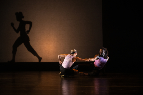 Two dancers on the floor reach right arms towards each other and connect. In the background, you see a dancer running behind the cyc.