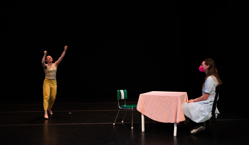 A solo dancer smiles with their arms outstretched high while an audience member sits at a table onstage, watching. A solo dancer smiles with their arms outstretched high while an audience member sits at a table onstage, watching.