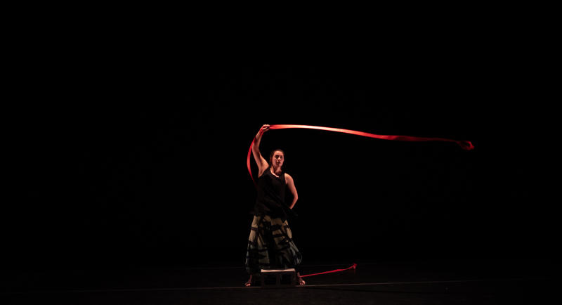 A solo dancer stands in front of a black background. They swing a long red ribbon. A solo dancer stands in front of a black background. They swing a long red ribbon.