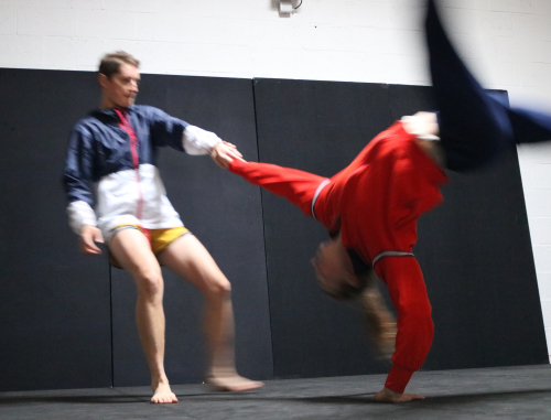 One dancer stands and one is upside down with one hand on the ground. They connect with one hand. 