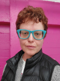 Headshot of the writer with turquoise rimmed glasses, standing in front of a bright pink wall.