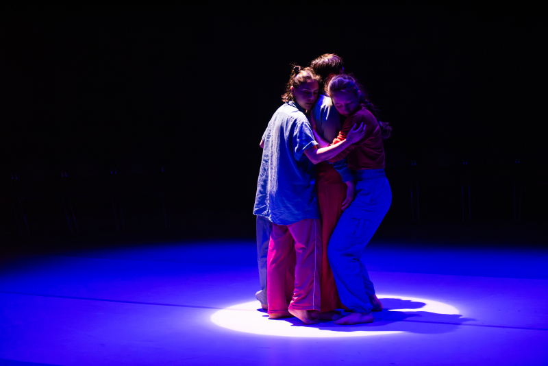     Four dancers clump together in a full-bodied group hug in a spotlight on an otherwise dark, blue stage.