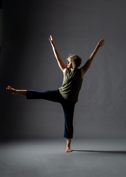 A dancer balances on her left foot, with her right leg lifted at hip level. Arms are up in a V shape.