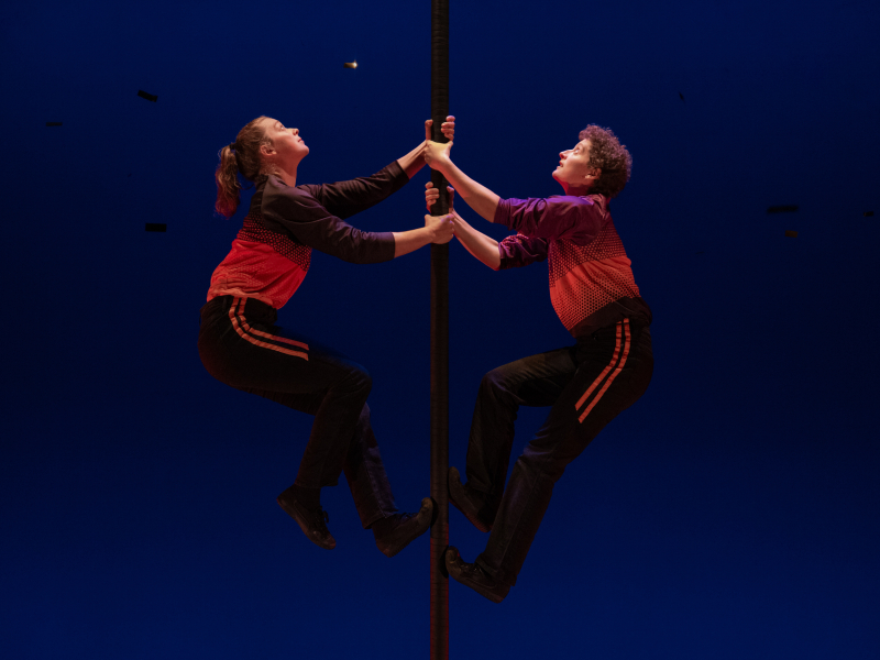 Two dancers hold onto a black pole and look up.