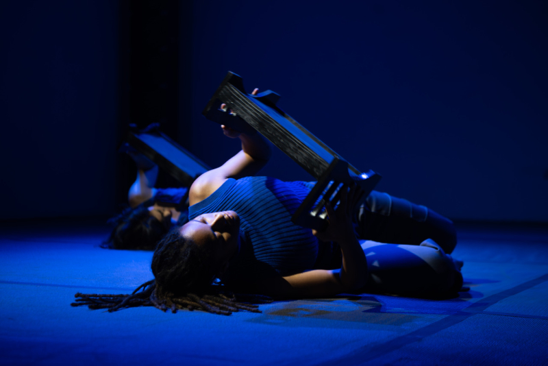 A dancer in blue light poses on the ground, holding a miniature bedframe.