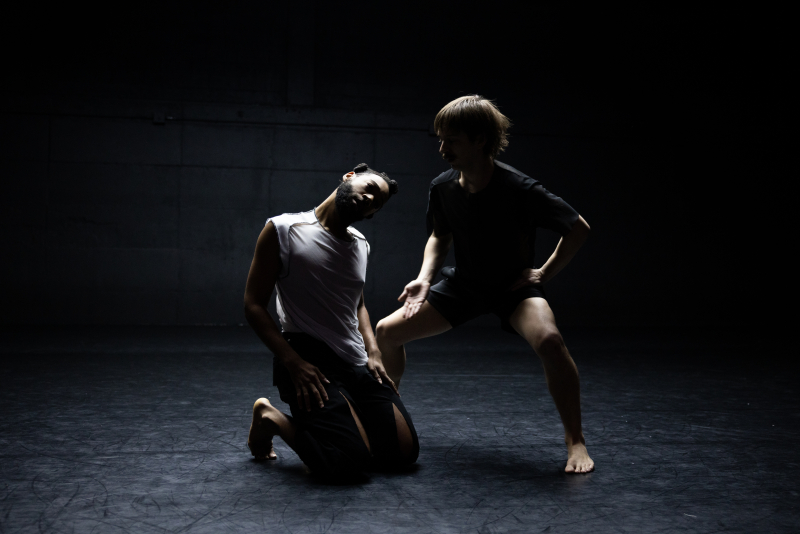 Two dancers dimly lit, one on their knees and one nearby in a wide second position plié.