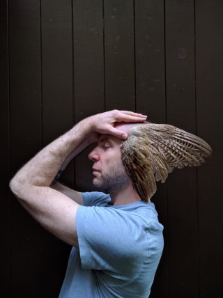 A headshot image of a male dancer facing right, holding bird wings/feathers by his ears.